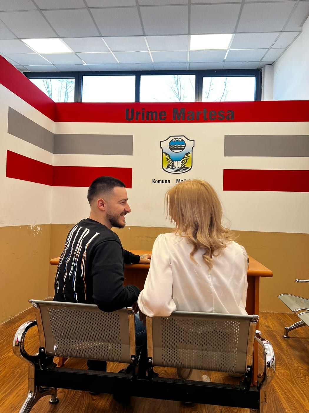 Rifat und Ela sitzen gemeinsam unter einem Schild mit der Aufschrift Urime Martesa in der Gemeinde Malisheva