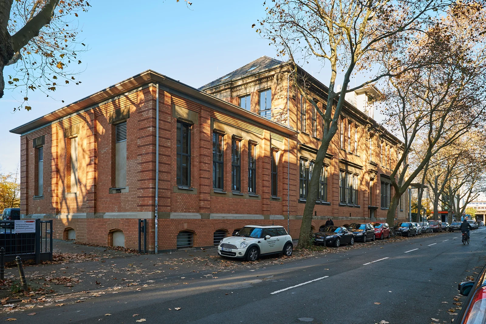 The brick exterior of Filmhaus Köln on Maybachstraße, with autumn trees, parked cars and a cyclist.