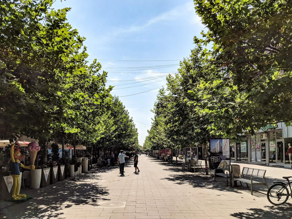 Ferizaj'ın merkezinden geçen ağaçlı yürüyüş sokağı; gölgede dükkân cepheleri ve kafe masaları