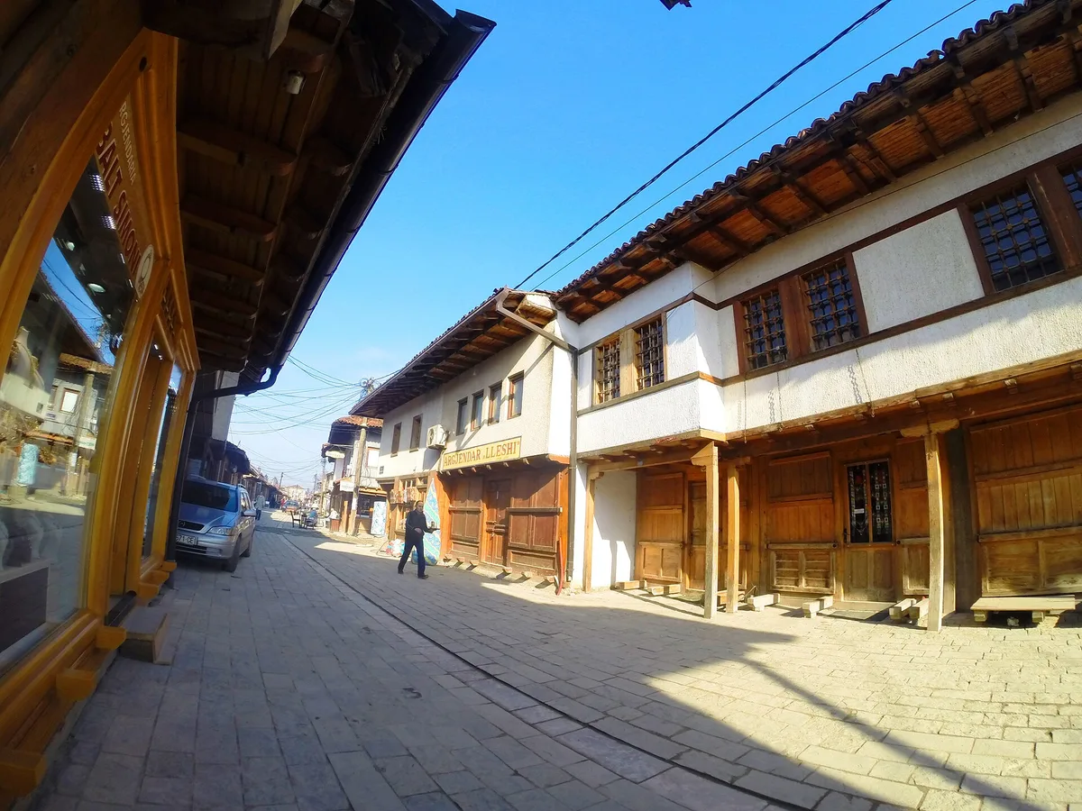 Gjakova'daki Büyük Çarşı'nın taş döşeli bir sokağı boyunca, çıkma kiremitli saçaklı ahşap dükkân cepheleri