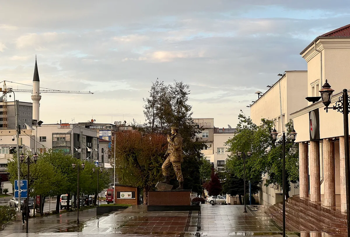 Yağmurlu bir akşamda Gjilan'ın yürüyüş merkezi; meydanın ortasında kaide üzerinde bronz bir heykel, arkada bir minare ve bir inşaat vinci