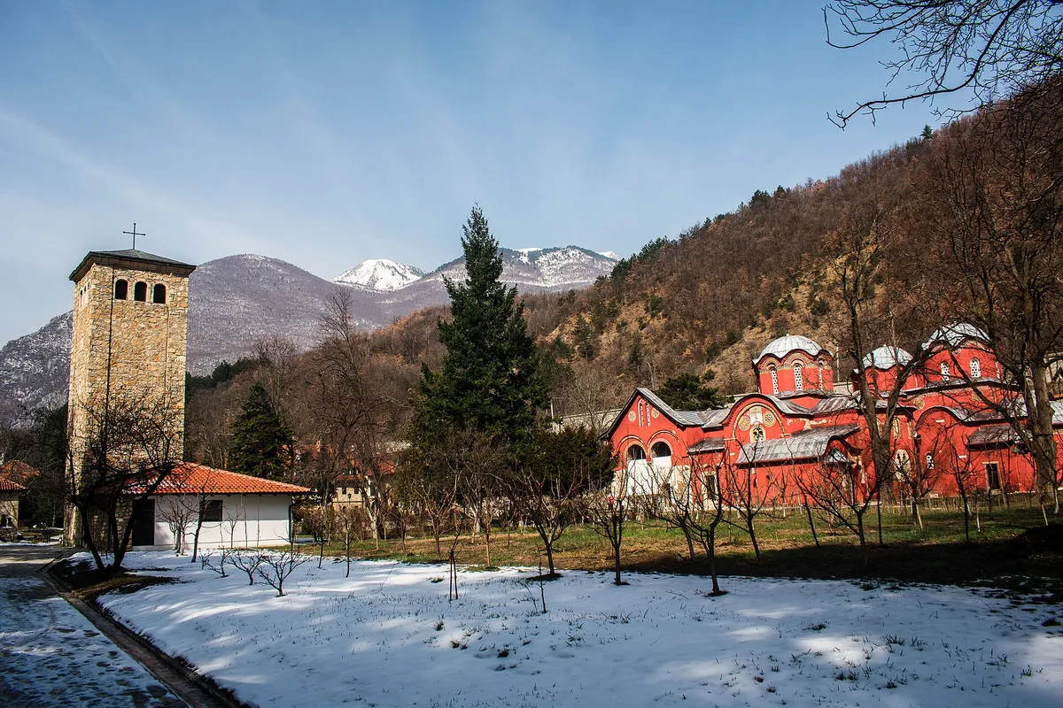 Peć Patrikhanesi'nin kurşun kubbeli, kırmızıya boyanmış kilise topluluğu; yanında taş bir çan kulesi, yerde kar, arkada dağlar