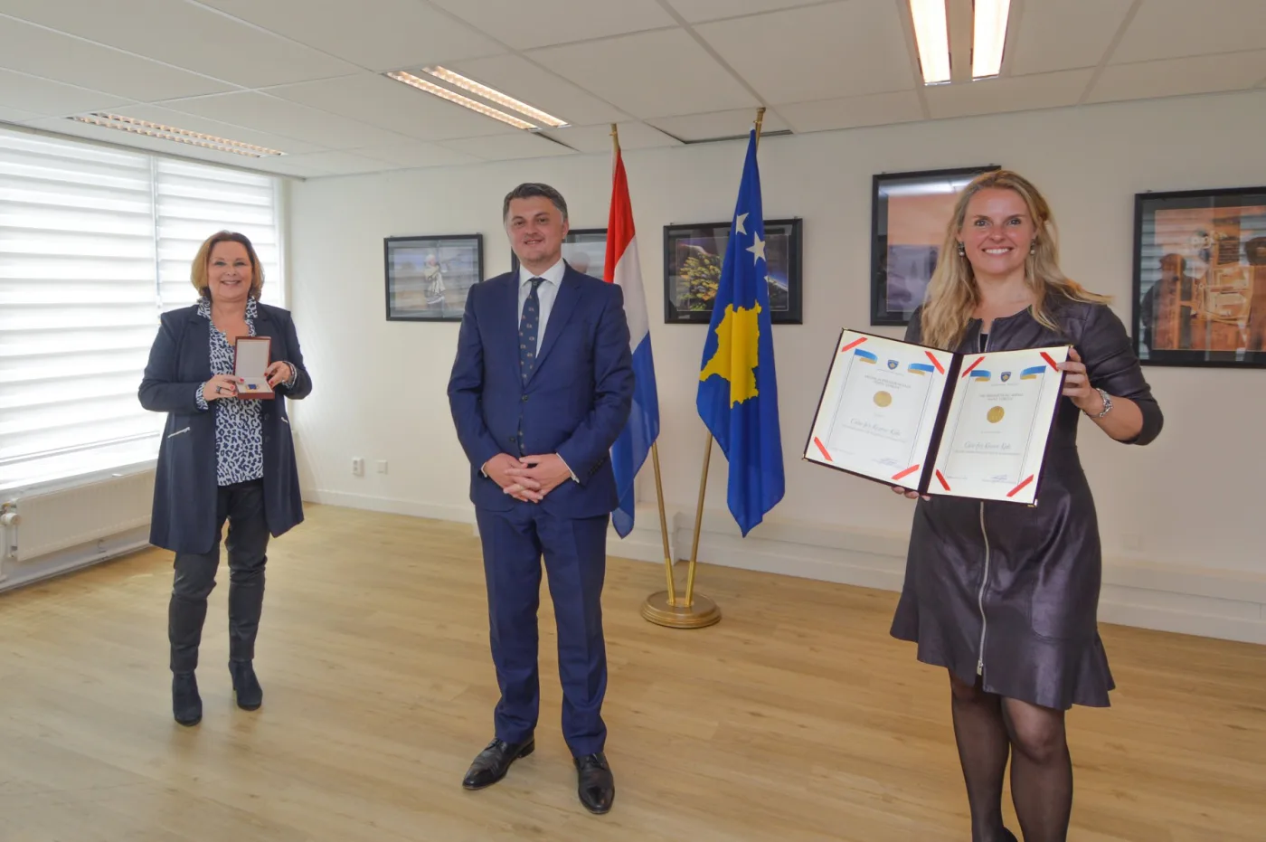 El embajador de Kosovo en La Haya entrega la Medalla Humanitaria Madre Teresa a dos voluntarios neerlandeses de Care for Kosovo Kids, con las banderas neerlandesa y kosovar detrás