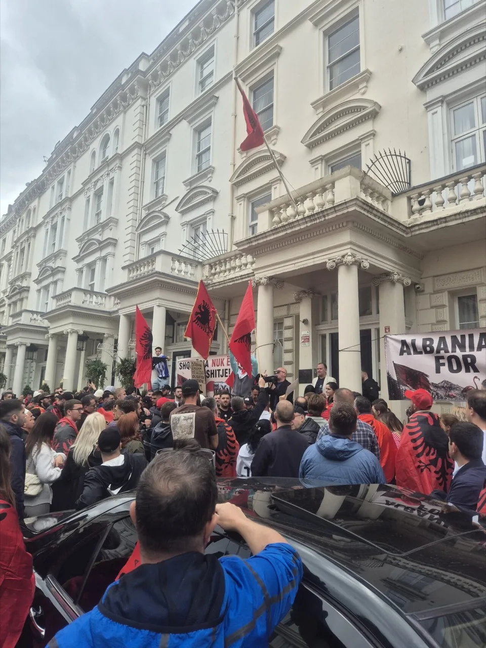 Eine Menschenmenge mit rot-schwarzen albanischen Flaggen und Transparenten vor der albanischen Botschaft in London, einer weiß verputzten Terrasse