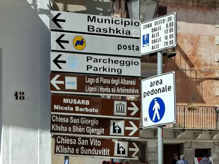 Un conjunto de señales en Piana degli Albanesi indica el ayuntamiento, el lago y dos iglesias en italiano y arbërisht