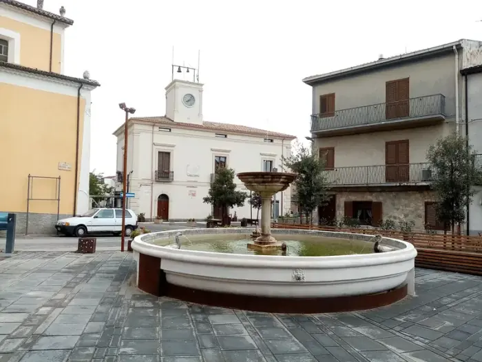 Piazza Skanderbeg in San Basile mit einem runden Brunnen und einem weißen Gebäude mit Uhrturm, der die Jahreszahl 1914 trägt