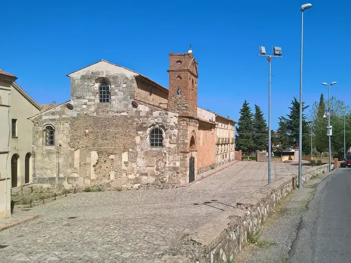 La antigua iglesia de piedra de Sant’Adriano, con campanario de ladrillo rojo y plaza empedrada, en San Demetrio Corone bajo un cielo azul