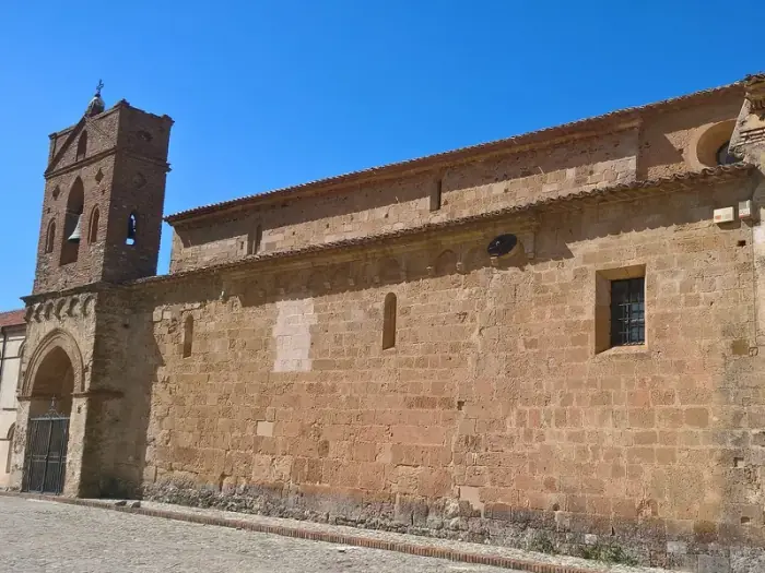 El largo costado de piedra de la antigua abadía de Sant’Adriano en San Demetrio Corone, con su campanario de ladrillo, bajo un cielo azul