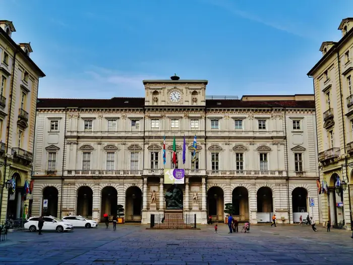 La fachada de piedra clara del Palazzo Civico de Turín, con su torre de reloj y banderas, vista desde una plaza pavimentada