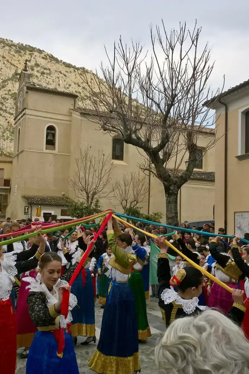 Junge Frauen in roten, blauen und grünen Trachten tanzen bei den Vallje auf dem Platz von Civita mit langen bunten Bändern unterhalb eines Kalksteinfelsens