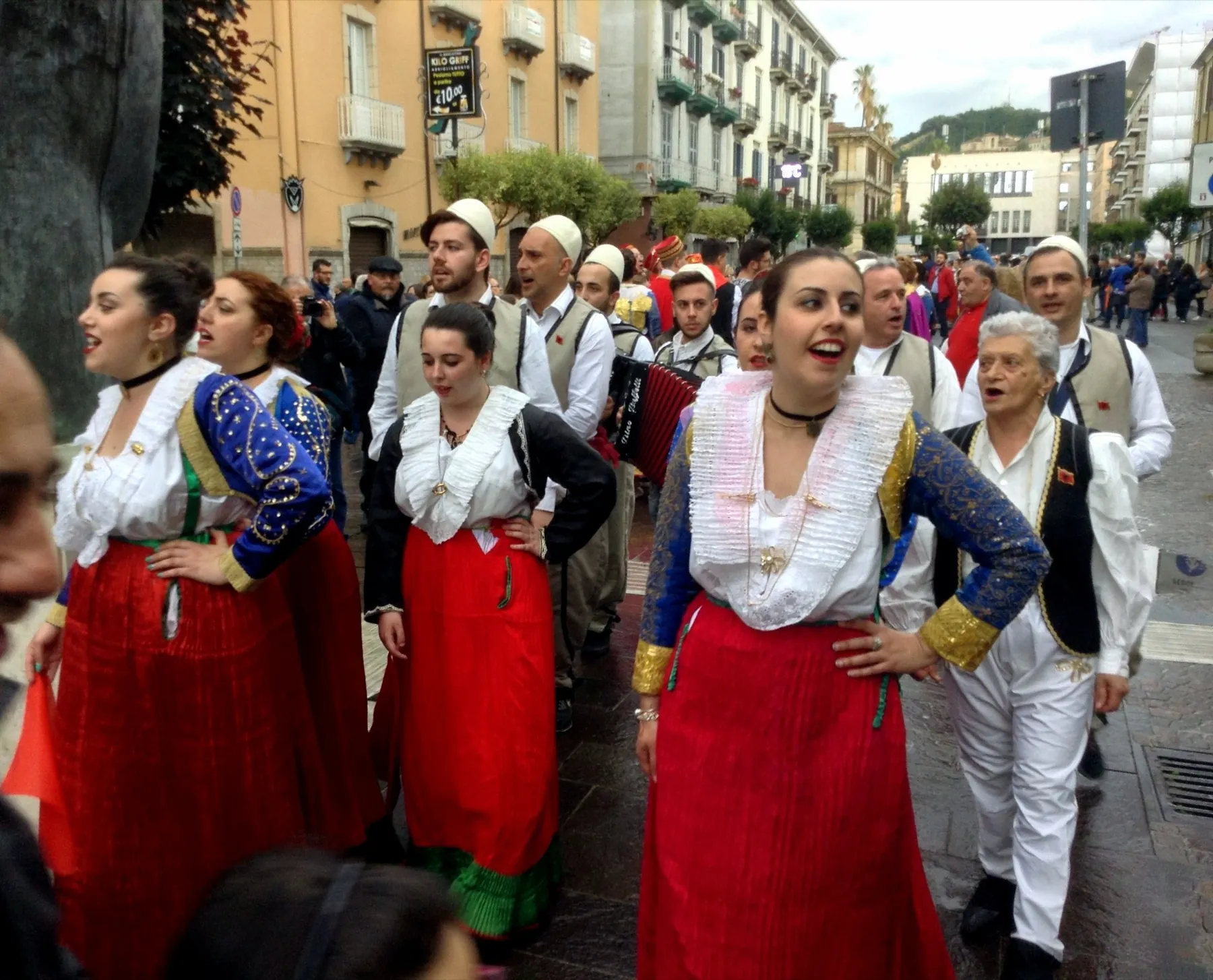 Jóvenes de Lungro con traje tradicional cantan con las manos en las caderas, mientras detrás cantan hombres con gorros blancos de fieltro