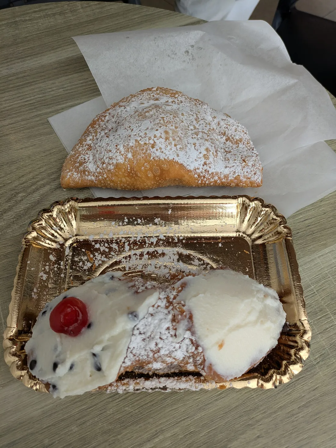 Un cannolo de ricota con una cereza confitada y una raviola frita azucarada sobre una bandeja de papel dorado