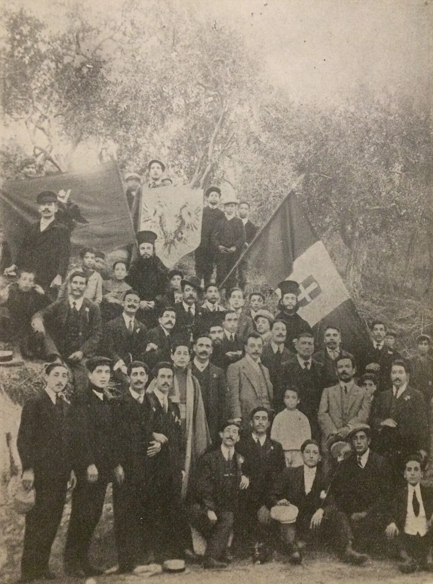 Fotografía de grupo en sepia de unos cincuenta hombres y niños con banderas bajo olivos en Piana degli Albanesi, 1911