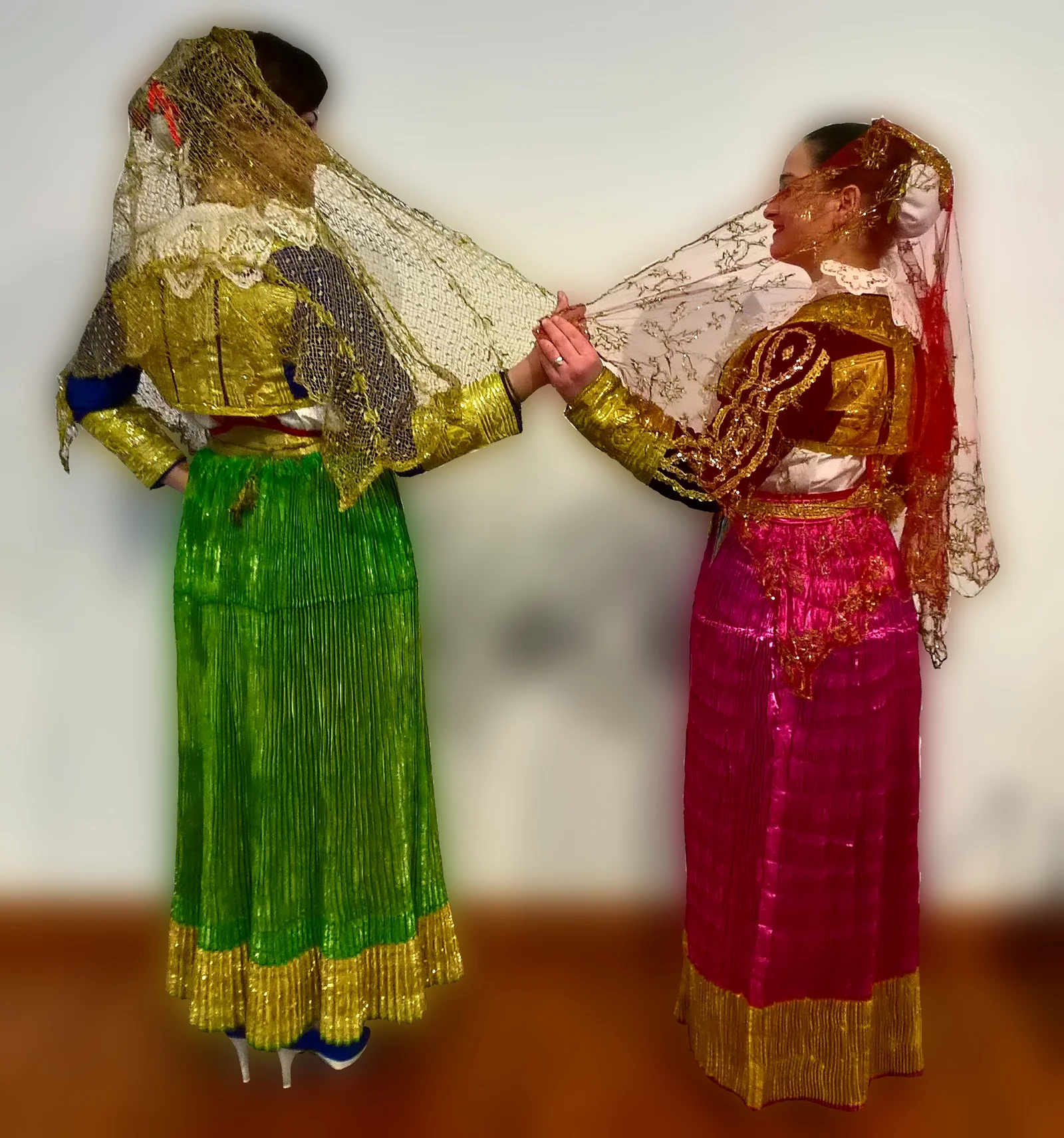 Dos mujeres con el traje de gala arbëresh de San Demetrio Corone, una de verde y oro y otra de magenta y oro, unidas por sus velos bordados en oro